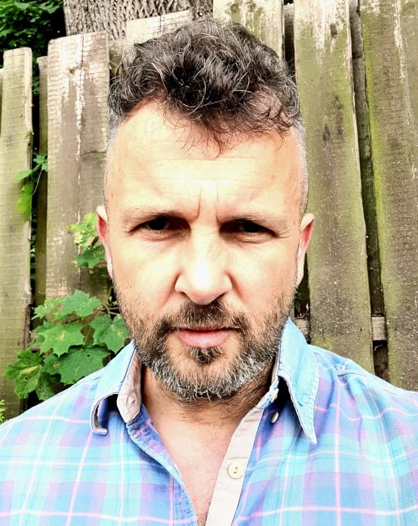 Bartosz Stanecki w koszuli w kratę na tle drewnianego płotu