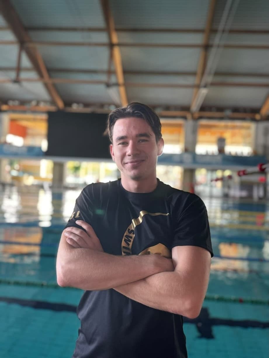 Jacek Michalski w czarnej koszulce Swimland ze skrzyżowanymi rękami na tle pływalni