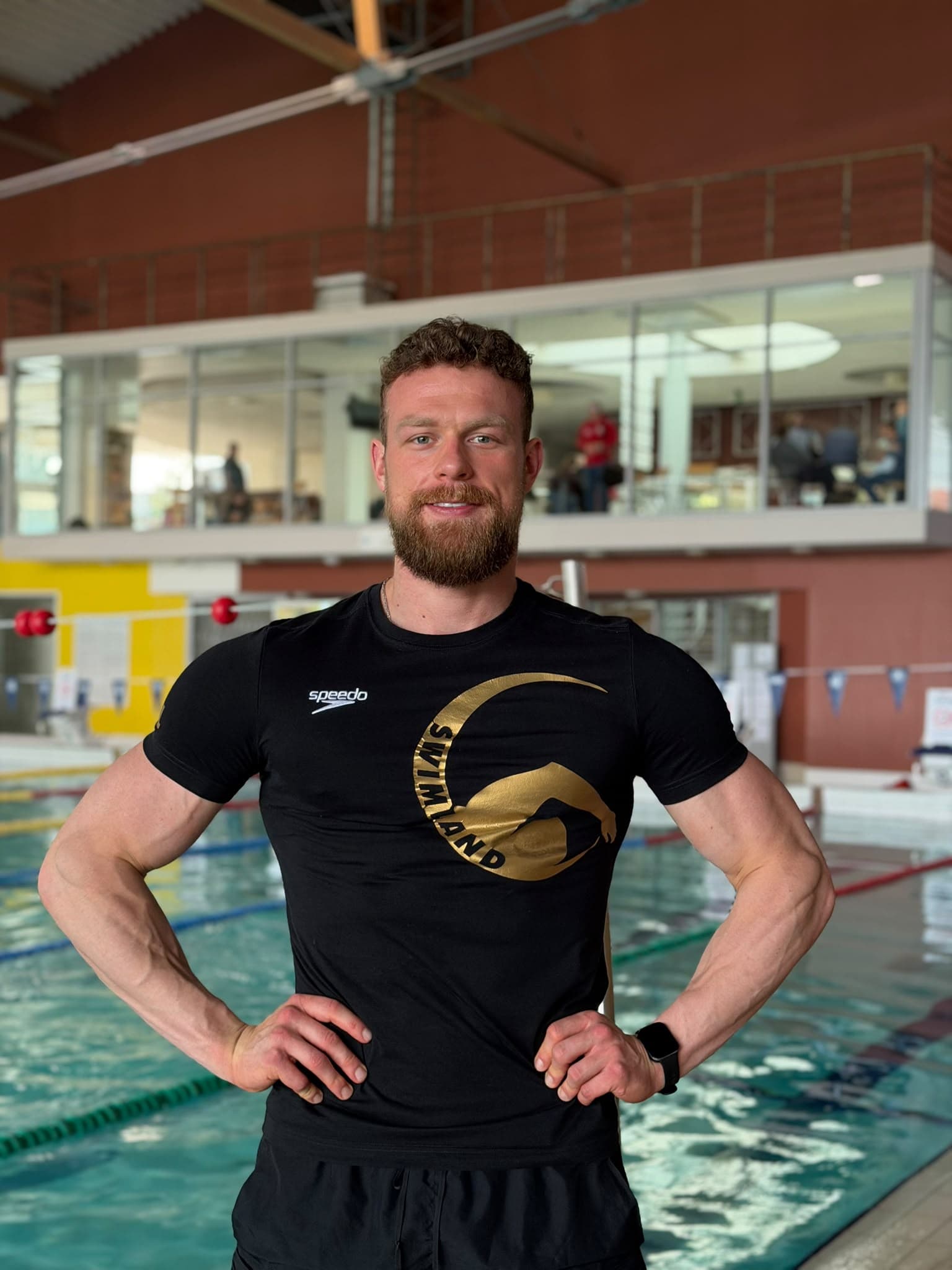 Karol Zbutowicz w czarnej koszulce Swimland z rękami na biodrach na tle pływalni