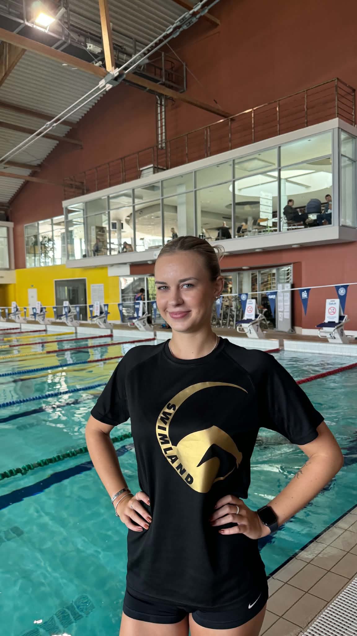 Oliwia Pustelak w czarnej koszulce Swimland z rękami na biodrach przy torach pływalni